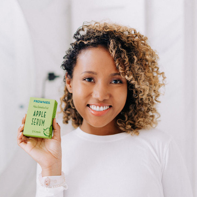 A black woman with curly hair shown holding Frownies apple serum in her right hand while smiling 