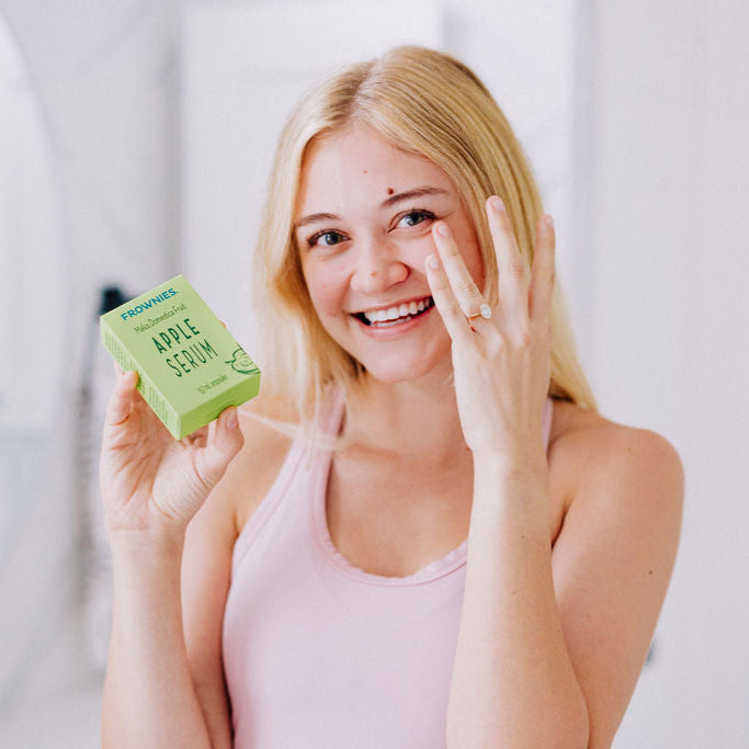 A white woman with blonde hair shown holding apple serum in her right hand while applying some to her left under eye area with her left hand 