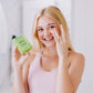 A white woman with blonde hair shown holding apple serum in her right hand while applying some to her left under eye area with her left hand 