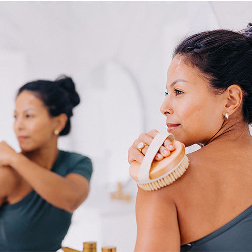 woman with dark hair pulled into a bun looking in mirror and using round body brush to massage shoulder 