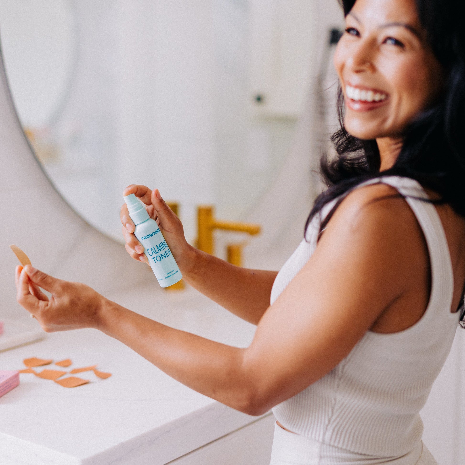 A woman smiling and using Frownies calming toner to wet her Frownies facial patch before applying it 