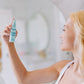 A woman with blonde hair smiling and holding Frownies calming toner up as she is bout to spay it onto her face 