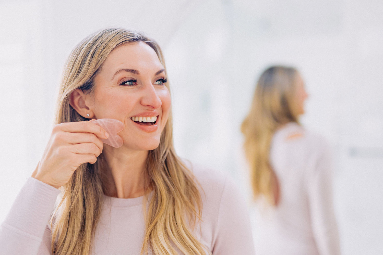 woman with long blonde hair wearing light pink long sleeved shirt performing facial massage with rose quartz stone 