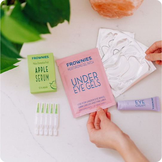 frownies apple serum, eye cream, and eye gels laid out on a bathroom counter with woman's hands