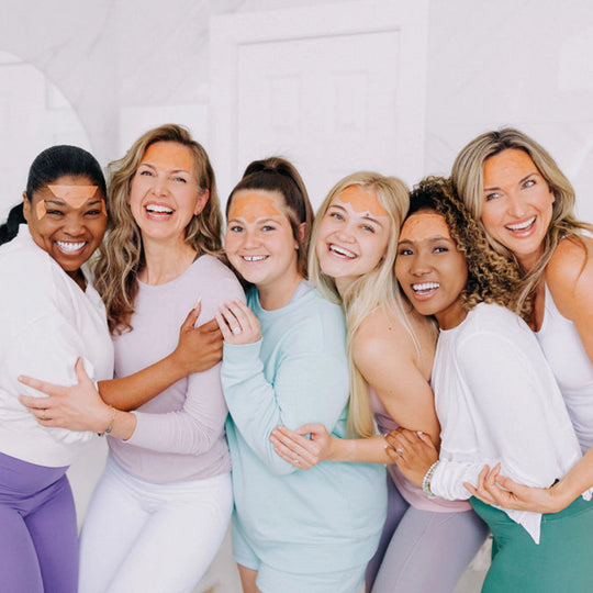 A group of six women close together, smiling and enjoying themselves while wearing Frownies facial patches.