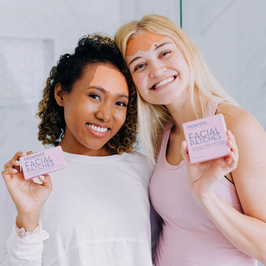 Two women smiling with their shoulders touching, wearing Frownies facial patches while holding the packaging up
