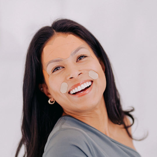 Close-up of a woman with black hair smiling while wearing Frownies Gentle Lifts, showing her shoulders and upper body.