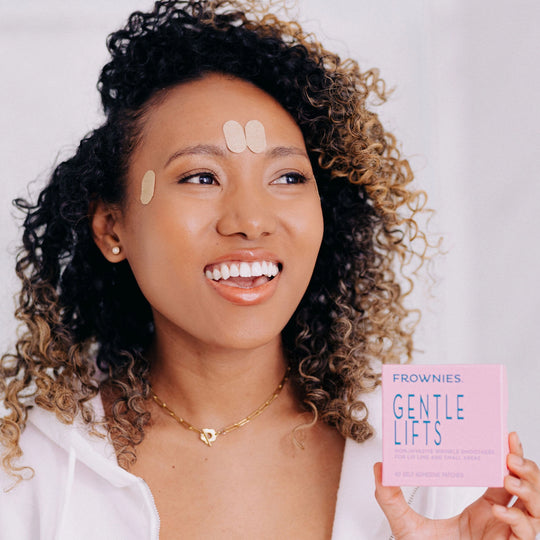 A Black woman with curly hair smiling while wearing Frownies Gentle Lifts and holding the box up.