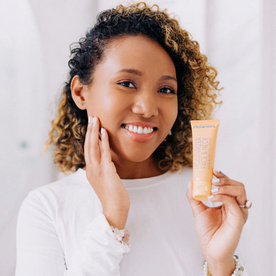 A curly-haired Black woman smiling, gracefully touching her face with her right hand while holding Frownies moisturizer for face and neck in her left hand.