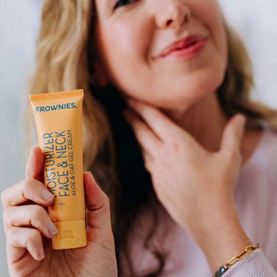 A close-up of a woman with a blurry face, holding Frownies moisturizer for face and neck in her right hand.