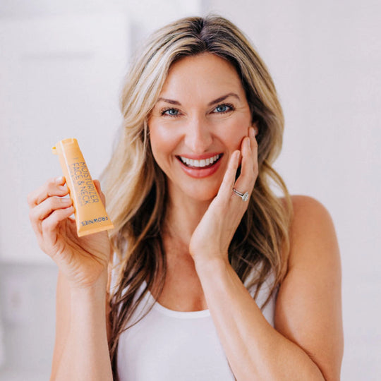 A blonde woman smiling, gracefully touching her face with her left hand while holding Frownies moisturizer for face and neck in her right hand.